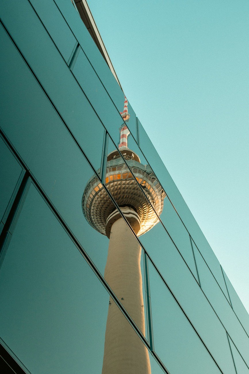 A television tower reflected in the glass facade of another building, the two structures overlapping